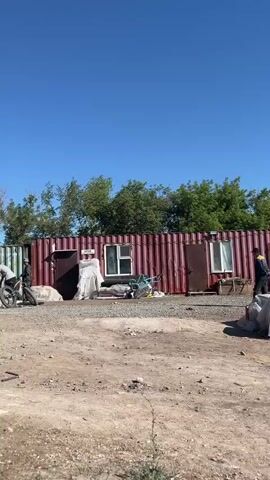 The video shows an outdoor scene with several people gathered around a structure made of shipping containers. The ground is dirt and gravel. Trees are visible in the background, and the sky is clear and blue.

In the foreground, a person on a bicycle is partially visible.

In the middle ground, near the shipping containers, several individuals are present. Some appear to be working or interacting with equipment. There are metal objects, possibly pipes or scaffolding, and what look like welding tanks. One person is seated on a makeshift step structure, and others are standing or crouching.

The shipping containers appear to be adapted for some use, with doors and windows visible. One container on the right has a window and some white sheeting or tarpaulin.

The overall impression is of an industrial or workshop setting, possibly under construction or in use for some kind of fabrication or repair work. The people are dressed casually, and the atmosphere seems relatively calm.