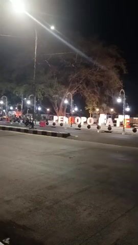 At nighttime, a wide, paved road runs in front of a low-rise building with white walls and a dark roof. Large, illuminated three-dimensional letters spelling out "PENDOPO BATE ALOS" are arranged in a semicircle on a dark, paved area to the left of the road. The letters are white with some of them illuminated in red. Several spherical, dark-colored objects are placed between the letters. In the background, behind the letters, are some trees, streetlights with lamps, and a tall antenna. Several people are visible in the scene, including one person riding a motorcycle from left to right across the frame in front of the letters. Another person is standing near the letters. The building in the background appears to be illuminated from within, and people are visible inside its entrance. The overall lighting of the scene is dim, with the illuminated letters and building providing the primary light sources in the darkness.