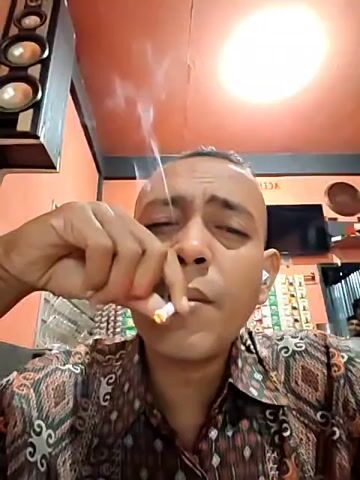 Here is a description of the video:

The video shows a close-up of a man smoking a cigarette. He has a shaved head and is wearing a patterned shirt. He appears to be indoors in a room with orange and gray walls. Speakers are on the upper left and a TV is behind him. A window covered in vertical blinds is on the left side. The man takes a drag from the cigarette and exhales a large plume of smoke into the air. Another person is visible behind him.