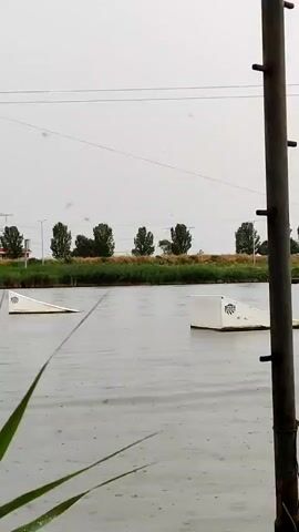 This video captures a wakeboarder in action on a calm body of water, likely a cable park, given the presence of ramps and a pulley system visible in the background. The scene is set against a backdrop of trees and a slightly overcast sky, suggesting it's daytime, possibly late morning or afternoon. The water is disturbed by the wakeboarder's movement, creating a spray as they glide across the surface. The rider, dressed in a red vest and blue helmet, is seen performing a trick, launching off a ramp and airborne before landing back on the water. The overall atmosphere appears to be one of athletic pursuit and enjoyment, with the weather being neutral, possibly a bit cloudy. The presence of reeds in the foreground adds a natural element to the otherwise man-made wake park environment. There are no discernible landmarks or text that specifically pinpoint the location within Honselersdijk, Netherlands, beyond what is implied by the general scenery of a European freshwater lake or canal. The focus remains on the wakeboarder's skill and the dynamic interaction with the water.