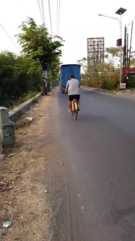 A person on a bicycle, wearing a light-colored shirt and dark pants, rides down the right side of a paved road, moving away from the camera. In front of the cyclist, a large blue truck travels in the same direction. To the right of the cyclist, and further ahead, a person on a motorcycle, wearing dark clothing and a helmet, rides in the same direction. The road is wet, suggesting recent rain. To the left of the road is a dry, grassy shoulder with some debris, bordered by a low concrete wall. Beyond the wall, there is dense green foliage and trees. Overhead, power lines stretch across the sky. In the background, on the right side of the road, a tall billboard-like structure is visible, along with streetlights and more trees and buildings. The sky is overcast.