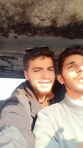 The video shows three young men inside a vehicle. The first part of the video shows two of them in the front seat. The first man has dark hair and a beard and is wearing a black jacket and tan shirt. The second man is next to him and is wearing a gray sweater. The camera then shifts to show the third person in the back seat, a woman with reddish hair, wearing a black and white plaid scarf. The interior of the car appears worn.