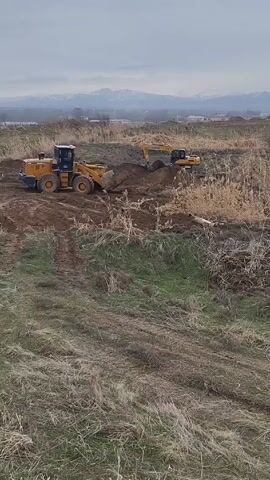 The video captures a scene of earthmoving activity. A yellow wheel loader sits on a dirt mound on the left side of the frame, its bucket near the ground. In the background, slightly to the right, an excavator is actively digging into the soil. The surrounding terrain is a mix of dirt, dry grass, and some green vegetation. A few dead tree branches lie on the ground. In the far distance, a mountain range is visible under a cloudy sky. The camera appears to be stationary, capturing a wide shot of the ongoing construction or land clearing work.