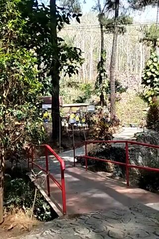 A narrow footbridge with red railings spans a small ditch. The bridge is made of concrete and is surrounded by lush greenery. In the background, there are a few small buildings with people visible inside and around them. Some people are wearing yellow vests. The area appears to be a park or recreational area with trees and a hilly terrain. The sun is shining, creating shadows.