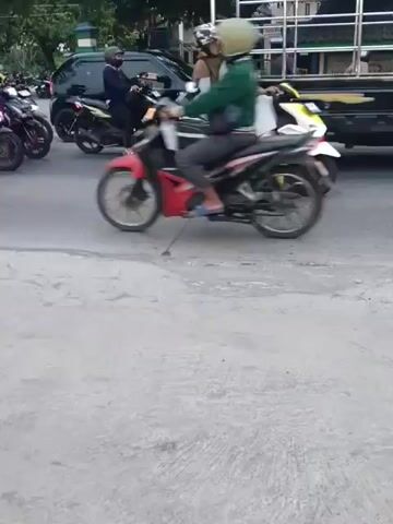 The video shows a street scene with multiple vehicles. In the foreground, there is a gray asphalt road with some cracks. In the middle ground, there are several motorcycles and cars. One motorcycle with two riders, one wearing a light-colored hijab, passes by. Another motorcycle with a single rider in a dark jacket is also visible. Further in the background, a black SUV and a green off-road vehicle are seen. The environment appears to be a street in a town or city, with buildings and trees lining the road. The lighting suggests it might be daytime, possibly overcast.