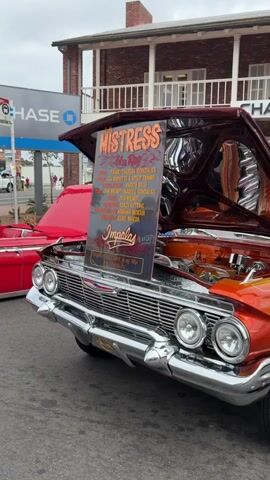 A classic orange and yellow lowrider car is parked outside a Chase bank. The car's hood is open, revealing a chrome engine. A sign stands in front of the car's grille, advertising "Mistress" and listing names. A man in a straw hat is standing next to the open trunk of the car, holding a blue and purple object. Tents are set up behind the car. The sky is overcast.