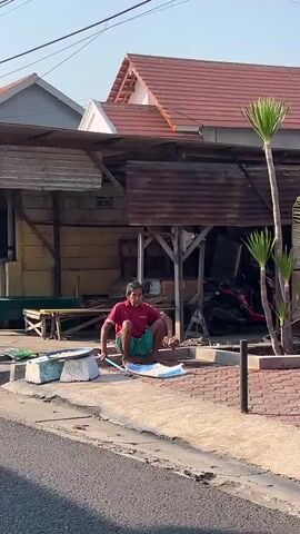 A man in a red shirt and green shorts is squatting on the sidewalk in front of a building, using a broom and dustpan to sweep debris into the dustpan. He is facing away from the camera and appears to be focused on his task. The building behind him is made of wood and corrugated metal, and it appears to be a small shop or market. There is a tree with large, fan-shaped leaves to the right of the man, and a motorcycle is parked behind the building. The sun is shining, casting shadows on the ground, suggesting it is daytime.