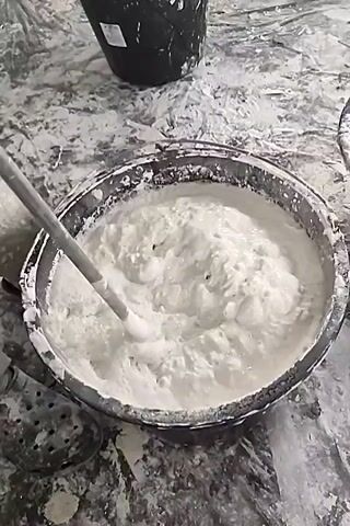 The video shows a close-up of a dark gray tub filled with a thick, white, frothy mixture, likely plaster or a similar building material. A long, light-colored mixing stick is submerged in the mixture, actively stirring it. The mixture is churning and splashing, creating bubbles and a creamy, somewhat lumpy texture.

The tub is situated on a surface that appears to be covered in the same white material, suggesting a work area where construction or renovation is taking place. Some of the white material has splashed onto the sides of the tub and the surrounding surface, indicating vigorous activity. In the background, a portion of a black bucket is visible. The lighting is somewhat dim, but it appears to be daytime. There are no people clearly visible in the frame, though the presence of the mixing stick implies human involvement.

The overall scene depicts a preparatory stage of a construction or DIY project, specifically the mixing of a plastering compound. The focus on the material and the mixing action suggests the importance of achieving the correct consistency for application. The environment appears utilitarian and industrial. No visible text is present in the video.