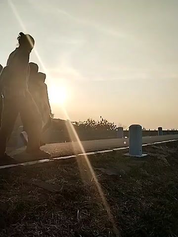 This video captures an outdoor scene in Jambeyan, Indonesia, during either sunrise or sunset, characterized by warm, low-angle light that silhouettes the subjects.

The footage opens with two individuals walking away from the camera on a paved path. The person on the right, with hair tied up and wearing a backpack, is slightly in front of the other. Shortly after, a third person joins the group, and then a fourth, forming a line of four individuals walking in the same direction. All figures are rendered in silhouette against the bright, golden sun, which is prominently visible on the left side of the frame, creating a distinct lens flare.

The setting appears to be an elevated path, possibly a dike or embankment, with a dry, grassy, and dirt verge alongside it. Blue and white bollards are visible at regular intervals along the edge of this verge. In the background, distant trees and foliage are outlined against a hazy sky, completing the natural landscape. The weather is clear and sunny.

The primary activity depicted is a leisurely walk, suggesting a recreational or exercise outing. Due to the silhouetted nature of the subjects, their expressions and emotions are not discernible. No visible text is present in the video.