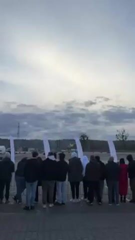 The video captures a group of people standing behind a low barrier, looking towards a yellow helicopter taking off from a paved area. There are multiple white banners with a logo, and a tall metal tower in the background, as well as trees. The sky is filled with gray clouds. One person has a child on their shoulders for a better view. The helicopter is the focal point and is seen ascending against the cloudy sky. The people are diverse in their attire, mostly wearing casual clothing suitable for an outdoor event.
