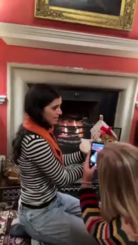 Two women are in a room with a fireplace. One woman with long dark hair, wearing a striped shirt, jeans, and an orange scarf, is kneeling in front of the fireplace, holding up a small gingerbread house on a white platform. The fireplace is lit, and there is a decorative log holder to the left. The second woman, with long blonde hair and wearing a multicolored striped sweater, is standing and taking a photo of the gingerbread house and the woman holding it with her smartphone. The walls are red, and there's a painting above the fireplace. A patterned rug is on the floor. The woman kneeling moves the gingerbread house around for the photograph.