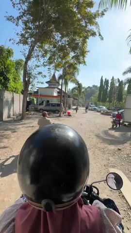 The video begins from the perspective of someone riding on the back of a motorcycle, looking forward over the driver's helmet. The motorcycle is on a dirt road with trees on either side. In the distance, there is a building with an orange roof and white walls, possibly a mosque. There are several parked cars and other motorcycles on the right side of the road. A person is seen walking in the distance. The sky is clear and blue. The motorcycle then begins to move forward.
