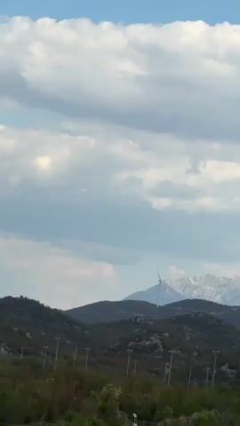 The video shows a scenic landscape with a highway in the foreground and mountains in the background. Several cars are driving on the highway, and there are wind turbines visible on the mountains. The sky is cloudy, and the overall impression is one of a beautiful, natural setting with a touch of modern technology. The video is shot from a moving vehicle, likely the passenger seat of a car, as the camera pans across the landscape. The perspective is from a slightly elevated position, offering a clear view of the road and the surrounding environment. The trees and bushes on the side of the road are lush and green, indicating a temperate climate. The mountains are covered with a mix of trees and rocks, and some of them have patches of snow at their peaks. The wind turbines are tall and slender, with large blades that are slowly rotating. The highway is well-maintained and appears to be a major route, with multiple lanes of traffic. The cars on the road are a mix of sedans and SUVs, and they are all moving at a moderate speed. The lighting in the video is somewhat dim, suggesting that it may be late afternoon or early morning, or that the sky is heavily overcast. The clouds are a mix of white and gray, and they are casting shadows on the landscape. There are also several streetlights visible along the highway, which are spaced at regular intervals. The overall mood of the video is peaceful and serene, with a sense of movement and travel.