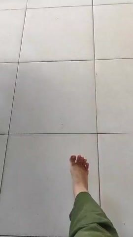 A pair of bare feet is shown walking on a tiled floor. The camera appears to be mounted at eye level, looking down at the feet and the floor. The tiles are a light color, possibly white or cream, and are laid in a grid pattern. The feet are positioned in the bottom portion of the frame, and as the video progresses, they move further up and to the right. The movement suggests the person is walking away from the camera. The lighting is even, and there are no other objects or people visible in the frame. The overall scene is simple and focused on the act of walking.