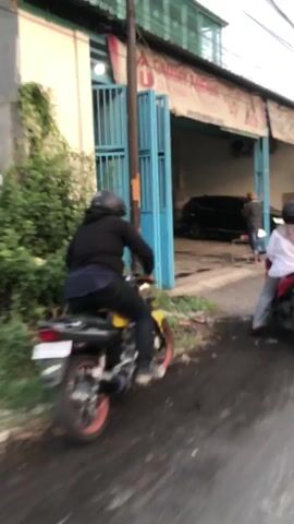 The video shows a street scene from a moving perspective, likely from a vehicle. In the foreground, a person on a red and white scooter is moving away from the camera. The scooter rider is wearing a white hooded jacket and grey pants. The street is wet and appears to be paved with asphalt and some cobblestones. To the left, there is a building with a blue open garage-like entrance. Inside the building, two people are visible, and a motorcycle is parked. A black car is also parked in the garage. To the right of the garage, a building with a yellow section and some red roofing is visible. A woman in a green dress is standing on the sidewalk to the right of the road. Overhead power lines are visible against the sky. The scene appears to be in an urban or semi-urban environment.