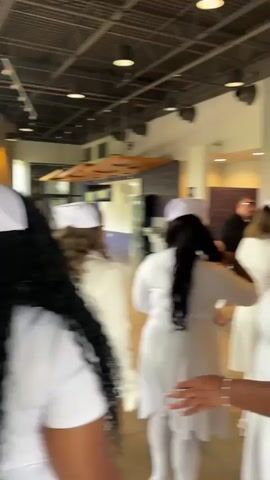 The video captures an indoor scene at a modern facility, likely a church or community center, identified by a large wall sign displaying "CROSSPOINT" and another sign partially visible as "worship ha" (suggesting "worship hall"). A large group of people is present, with many adults, primarily women, dressed in matching white uniforms and traditional white nurse caps. They appear to be lining up, mingling, and greeting each other, moving through a wide hallway or lobby area. Other individuals are dressed in various casual clothing, and some children are also visible. The setting features light-colored walls and flooring, with visible ceiling ducts and modern lighting fixtures. People appear cheerful and engaged, suggesting a social or celebratory event. The uniforms imply a specific organization or occasion, possibly a graduation, blessing, or special service for a healthcare-related or ministry group in Grand Prairie, United States of America. A banner asking "New Here?" is also visible.