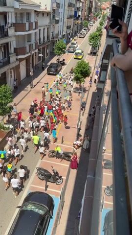 The video depicts an aerial view of a street event, possibly a race or parade, taking place on a sunny day. A large group of people, predominantly children and young adults, are seen running down the street. Many are wearing white t-shirts, while a smaller group is dressed in red. The street is lined with buildings, some of which have balconies. Several cars, including a police car, are parked or driving along the street. Motor scooters are also present. Spectators are visible on balconies and sidewalks, observing the event. The street itself is paved and marked with pedestrian crossings. The overall atmosphere appears lively and festive.