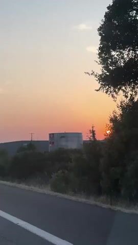 The video shows a view from inside a vehicle driving on a highway at sunset. The sky is a gradient of orange and pale yellow. In the foreground, the asphalt road with white lane markings is visible. On the right side of the road, there are trees and bushes. In the distance, there are industrial buildings and more trees. The sun is a bright orange orb low on the horizon, partially obscured by a building. Other vehicles can be seen on the highway. The camera appears to be mounted on the dashboard or windshield of the vehicle.