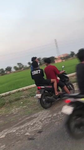Three young men are on a motorcycle. The motorcycle is black and has a license plate. The man in the back is wearing a black t-shirt with "1927" on it. The man in the middle is wearing a red t-shirt. The third man is partially visible on the right side of the frame. The motorcycle is on a dirt road next to a field of green crops. There are utility poles and trees in the background. The sky is overcast.