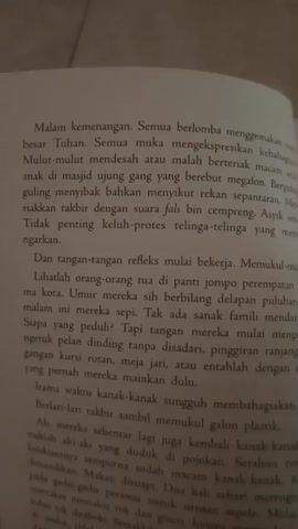 The video shows a close-up of an open book. The pages are filled with Indonesian text. The lighting is adequate, but the focus is slightly soft, suggesting the camera is not perfectly still or the lens is not optimized for this close-up. The book appears to be a novel or a collection of essays. The first page visible has a heading "Malam kemenangan." The second page has a heading "Maklum, hari raya." The text discusses themes of community, elderly care, and childhood innocence, possibly in the context of a religious holiday. There are no people visible in the video. The environment is indeterminate, but appears to be indoors, possibly a room with ambient lighting. The video is short, lasting about 5 seconds.