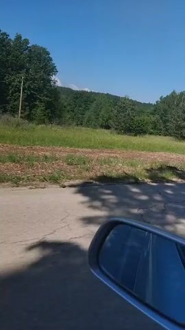 The video shows a drive through a rural area on a sunny day. The camera is positioned from inside a vehicle, showing a view from the driver's side mirror. A paved road runs through a landscape of green fields and trees. The sky is clear and blue. There are no visible people or interactions. The focus is on the scenery passing by the car. The video seems to be taken during the day.