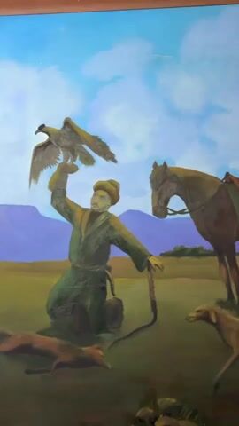 The video shows a painting of a falconer kneeling in a field, holding a falcon on his gloved hand. A horse stands to his right, and two dogs are in the foreground. The background features rolling hills and a sky with clouds. The falconer wears a traditional hat and green robes. One dog is walking, and the other is lying down. The horse is saddled and bridled. The scene depicts a hunting expedition.