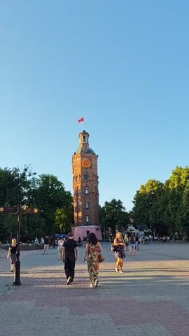 The video shows a wide shot of a public square with a distinctive clock tower as its focal point. The tower is made of brick and has a clock face with Roman numerals. A red flag flies from its top. Several people are walking across the square, which is paved with reddish-pink and grey bricks. The square is lined with trees on either side, and more people can be seen in the background, suggesting a lively atmosphere. The lighting indicates it is late afternoon or early evening, with long shadows cast across the square. The sky is clear and blue, suggesting good weather. The overall mood appears relaxed and pleasant, with people enjoying a stroll in the open space. The presence of the clock tower, a notable landmark, along with the public square setting, strongly suggests a location in a city or town center. Given the context of Vinnytsia, Ukraine, this is likely the Vinnytsia Water Tower, a historical monument and a popular tourist attraction. There is no visible text in the video.