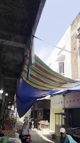 A vertical shot pans upwards along the exterior of a building with a brick facade partially covered by modern white siding. The building appears to be under construction or renovation, with an open, unfinished window opening on the top floor. Below the building's upper levels, a colorful striped tarp is stretched out, supported by a blue tarp that extends outwards, creating a makeshift awning or cover for the lower level. The blue tarp partially obscures storefronts below. Sunlight illuminates the scene, casting shadows and highlighting the textures of the brick and tarps.