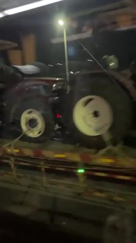 The video shows a dark, outdoor scene at night. The first frame shows a dark-colored tractor loaded onto a flatbed trailer. The tractor has large, white-rimmed wheels. The second frame is a closer view of the front of a white truck, possibly a transport vehicle, with reflective striping on the side. The third and fourth frames show the view from inside a vehicle, looking out at a road at night. A truck with its headlights on is visible in the distance, appearing to be moving. The headlights cast beams of light onto the road, and there is some motion blur, indicating the video is likely taken from a moving vehicle. The environment appears to be a highway or a well-lit road with barriers and streetlights.