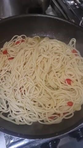 The video shows a close-up shot of spaghetti being prepared in a pan. The spaghetti is a light golden color, mixed with red cherry tomato halves and thinly sliced garlic. Fresh green parsley is being sprinkled over the top of the pasta. The pan is dark gray and appears to be on a stovetop, with some metallic elements visible in the background. The lighting is somewhat dim, focusing the attention on the food. The action of sprinkling the parsley is repeated throughout the short clip, suggesting the final touches are being added to the dish.