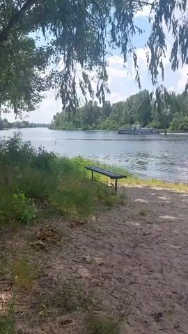 The video shows a body of water with a boat in the distance. In the foreground, there is a sandy beach with a wooden bench and some grass. A few children are playing near the water's edge. The sky is cloudy.