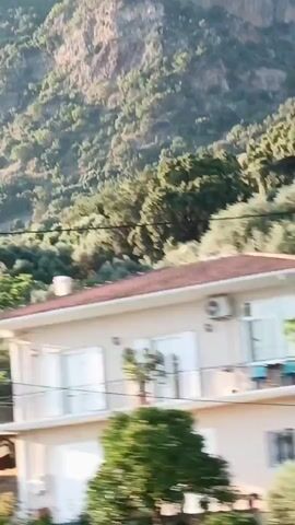 The video shows a view from a moving vehicle, looking out a window. The scene is a rural area with rolling hills covered in trees. In the foreground, there is a road with a guardrail. There are also some buildings and fences visible. The sky is clear and blue. The video is shaky, indicating that the vehicle is moving.