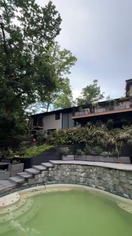 The camera pans up to show a two-story house with a pool in the foreground. The pool is oval-shaped and filled with green water. The pool's edge is made of gray stones. Above the pool is a wall made of similar stones, topped with a black wooden fence and several rectangular planters with various plants. The house is built into a hill. The first story is gray with black-framed windows. The second story is covered in dark wood and has a balcony with wooden railings and planters. Trees are seen on both sides of the house, with gray skies overhead.