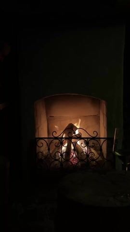 Here's a detailed description of the video:

The video shows a person adding a log to a roaring fire in a stone fireplace. The fireplace has a decorative black metal grate in front of it. The fire provides a warm, orange glow, illuminating the interior of the fireplace. To the right of the fireplace is a small, round wooden table, on which sits a partially filled wine glass. The person placing the log is wearing dark pants and a long-sleeved dark shirt. The walls of the room are a neutral shade, and additional firewood is stacked against the wall to the right of the fireplace. The scene is dimly lit, focusing the viewer's attention on the fireplace.