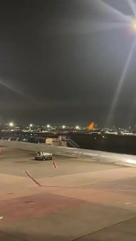The video shows an airport tarmac at night. An airplane wing is in the foreground, and the tail of a plane with an orange tip is visible in the background. The plane in the background appears to be on the tarmac or taxiing. There are lights from the city in the distance. A service vehicle is parked near the plane. The sky is dark and cloudy, with a bright light source visible in the upper right corner. There are no people clearly visible in the video. The overall atmosphere is dim and atmospheric, characteristic of an airport at night.