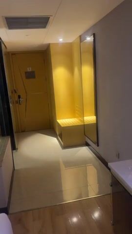 The video shows the interior of a hotel room. The room has a cream-colored tile floor and light walls with a dark gray accent wall. In the entry area is a golden yellow area with a seat built into the wall and a full-length mirror. The entry door is yellow. To the left of the bed, there is a partial glass wall that gives a view into the bathroom. The bathroom is lit with bright lights. A handbag with a brown and beige pattern and a green and red stripe is sitting on the white bed. There is a small bedside table next to the bed.