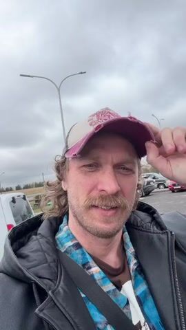 A man standing outdoors is speaking into a camera. He wears a distressed baseball cap, a blue and black plaid shirt, and a black leather jacket. He has light brown hair and a mustache. Behind him is a gas station with a green and white color scheme and the word "БЕЛНЕФТЕХИМ" is visible on the building. The sky is overcast and there are several vehicles parked in the background. The man mentions being in Belarus and heading to Lida. He says something about drinking Lida Kvas.