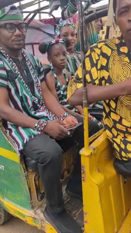 The video showcases a cultural celebration in what appears to be a small town. The initial scene focuses on a man in traditional attire sitting in the back of a yellow and green three-wheeled vehicle labeled "Kafanchan Mass Transit," alongside a child also dressed in traditional clothing. The man is wearing a green and black striped shirt, glasses, and a traditional cap. The video then shifts to a costumed figure adorned with layers of colorful straw-like material, moving along the street. The scene includes other individuals dressed in casual and traditional clothes, walking alongside the costumed figure. The video takes place in a public area, with buildings visible in the background.