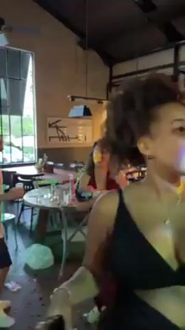 The video shows a woman in a maroon dress holding a baby, smiling at someone out of frame. The scene appears to be indoors at a celebration. Confetti litters the floor. Other people are visible, including the person holding the camera, who is wearing a black outfit.