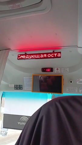 The video shows the interior of a bus, as seen from the perspective of a passenger. A digital display shows the bus stop name in Russian: "Я ОСТАНОВКА СВОБОДА", which translates to "I am the Freedom stop". Below the display, a small monitor shows a reflection, seemingly of the person recording. The temperature display reads 16°C. The bus's interior is white, with a blue tint on the windshield. A grey curtain is visible on the left side. The seat in front of the camera is occupied by a person whose head and shoulders are visible. The bus appears to be a YUTONG brand, as indicated by the logo on the windshield.