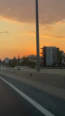 The video shows a sunset over a highway. A car is driving on the highway in the distance. There are buildings and trees in the background. The sky is orange and yellow with some clouds.