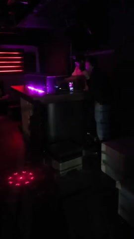 The video shows two scenes. The first is of a dark nightclub with a DJ booth lit in purple and red. Two people are standing near the booth, though details are unclear due to the darkness. The second scene transitions to an exterior shot at night, from an elevated position. A street with cars and motorcycles moving in both directions is visible, illuminated by streetlights and vehicle headlights. Buildings and trees line the street, and power lines are visible in the foreground. The overall lighting is low, with contrasting bright lights from the vehicles.
