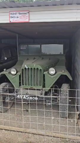 The video pans across several vintage vehicles parked under a covered structure. In the foreground, a green military-style jeep is visible, with large tires and a prominent grille. Behind it, a dark-colored sedan, likely a GAZ M-20 "Pobeda," is parked. The sedan is dusty and appears to be in a state of disrepair. The cover appears to be made of corrugated metal. A sign in Russian, partially visible at the top, indicates that the vehicles are from 1943 and 1948, and were made in the USSR. A wire fence is in front of the vehicles, suggesting they are on display in a museum or exhibition. The ground appears to be dirt and gravel. The lighting is overcast, suggesting it is daytime.