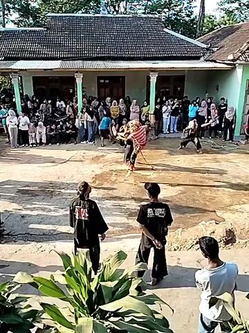 The video captures a scene in what appears to be a rural village. A traditional house with a tiled roof and columns stands in the background, with a crowd of spectators gathered in front of it. In the foreground, a dirt arena is set up for a performance. Two individuals in traditional martial arts attire are engaged in a dynamic demonstration. They are both wearing dark pants and tops with distinctive markings, and one is clad in an orange and brown patterned garment. Spectators, dressed in a variety of casual clothing, line the edges of the arena, observing the performance. Some individuals stand on the perimeter, possibly acting as organizers or participants waiting for their turn. The ground is uneven and dusty, indicative of an outdoor event. The atmosphere appears festive and engaged, with the onlookers focused on the martial arts display. The overall impression is of a cultural event or martial arts training session taking place in a community setting.