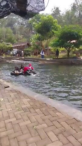 Three individuals, two women and one possibly a girl, are in an inflatable raft in a shallow body of water, possibly a river or canal. The woman in the red hijab is in the back of the raft, while the other two are in the front. The raft is moving downstream, and the people appear to be enjoying themselves. The water is clear and the bottom is visible. The banks of the waterway are lined with brick pavers and greenery. In the background, there are small buildings with thatched roofs and trees. Other people are visible in the background, some standing and some walking. The scene is well-lit and appears to be a sunny day.