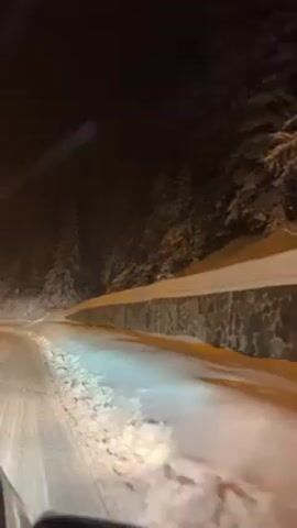 The video shows a car driving on a snow-covered road at night. The road curves to the left, with a low stone wall on the right side and snow-covered trees beyond. The car's headlights illuminate the road ahead, and snowflakes are visible in the air. The overall scene is dark and snowy.