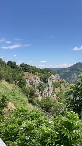 A vertical, outdoor video showcases a lush green mountainous landscape under a bright blue sky with scattered white clouds. In the foreground, dense green foliage with small white flowers obscures the bottom of the frame. The camera pans slightly to the right, revealing a steep, rocky cliff face covered in vegetation to the left. Further back, rolling green hills and mountains stretch into the distance, dotted with trees. A visible quarry scar or landslide is present on a mountainside in the mid-ground. A small village or settlement appears nestled in the valley below. The overall impression is one of a serene, natural environment. There are no people or discernible actions occurring.
