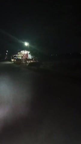 The video shows a nighttime scene from the perspective of a moving vehicle. A roadside stall is visible, illuminated by a streetlamp. The stall appears to be stocked with various items, possibly for sale. The road is paved, and there is darkness beyond the immediate vicinity of the stall and the road. Some blurred lights are visible in the distance. The overall environment suggests a rural or suburban setting at night.
