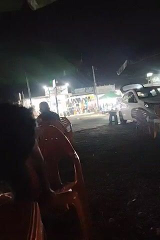 The video shows a scene at night. A young boy wearing a yellow Calvin Klein t-shirt is sitting on an orange plastic chair. He appears to be eating something. Other people, including a woman wearing a hijab, are seated nearby. The scene is under a green umbrella. A table in front holds plastic bags and a water bottle. Across a road are shops with bright lights. Motorcycles and a white car pass by in the background. The ground appears to be covered with small stones or gravel. The video seems to be taken from a low angle.
