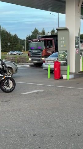 A motorcycle is parked at a gas station, facing left. To the right of the motorcycle, a silver sedan is parked next to a gas pump. A red gas canister is visible behind the sedan. A large red truck is parked behind the sedan, with its front bumper facing the camera. The truck appears to be a Scania. The gas station has a white canopy overhead. The sky is cloudy.